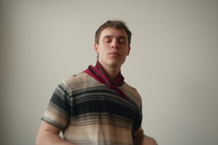 A man wearing a striped shirt and scarf sits against a plain wall adjusting the scarf with his hands