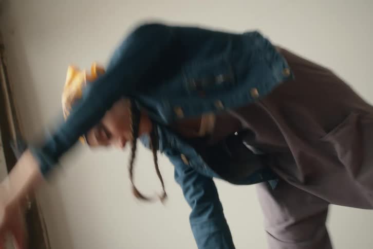 A woman wearing a headscarf denim jacket and pants is captured in motion with arms extended in a brightly lit space