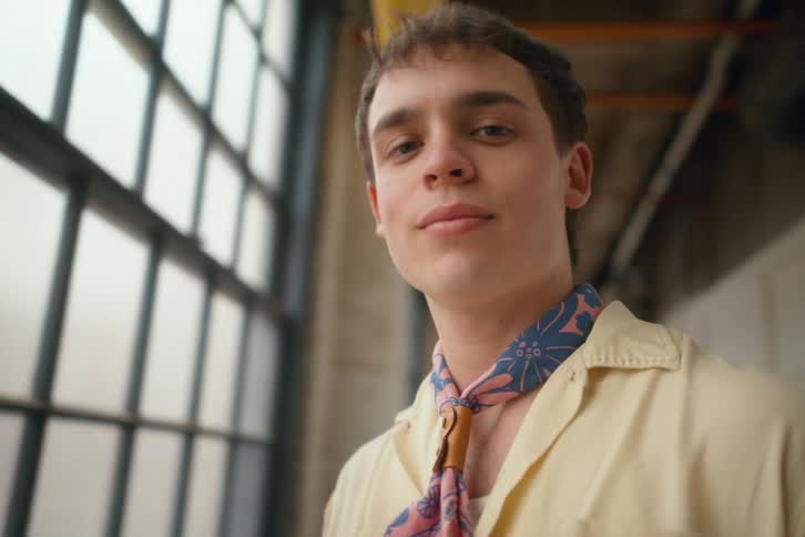 Male model smiles slightly as he strokes his shirt while standing in a long corridor near a large window