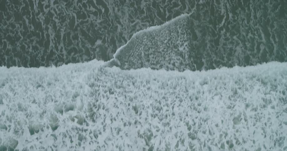 An aerial view captures the textured frothy surface of the ocean with white foam swirling against the deep green water creating a dynamic and abstract pattern near the sandy beach