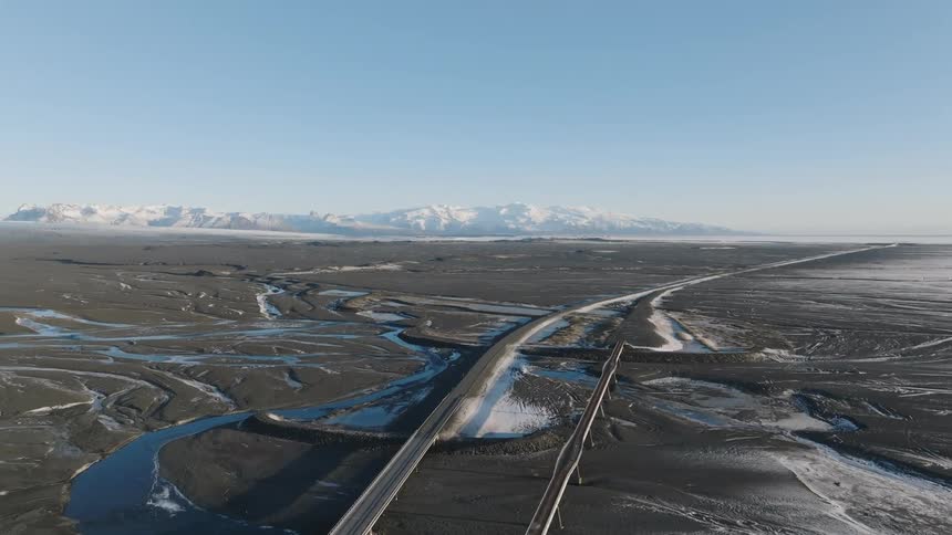 A pair of parallel bridges cross a braided glacial river plain in iceland stretching towards snowcovered mountains under a clear sky