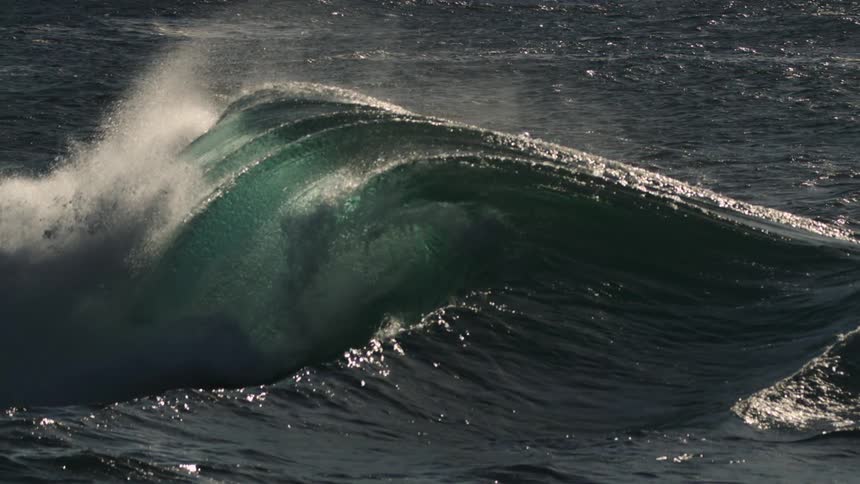 A dark rolling wave in the ocean its peak topped with white spray against the backdrop of a sunlit sparkling sea