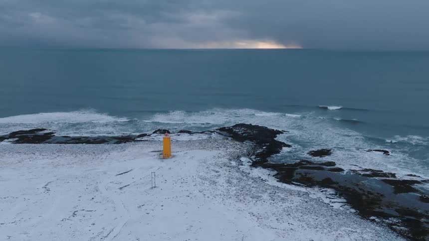 Waves crash onto a snowy rocky coastline that has a lighthouse