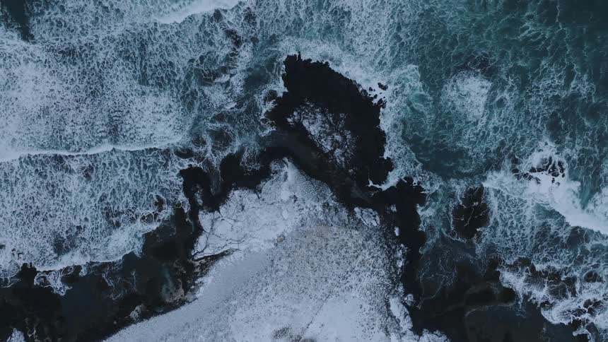 The dramatic meeting of icy land and turbulent ocean waves creating a striking contrast between the dark rocks and the frothy white surf