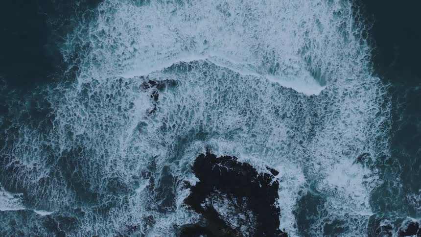 An aerial view of the textured frothy surface of the ocean with white foam swirling around dark jagged rocks creating a dynamic and abstract pattern that highlights the power and movement of the sea