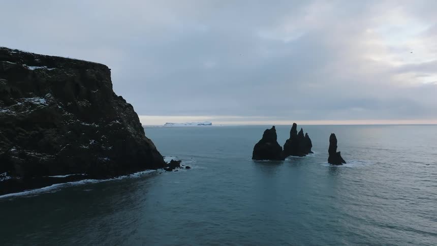 A dark rocky cliff jutting into the ocean with jagged rock formations emerging from the water against an overcast sky