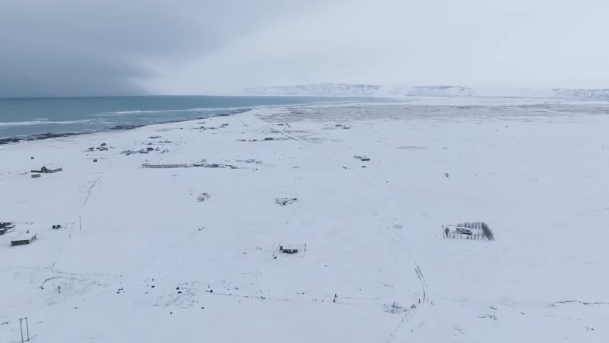 A snow covered coastal plain with small buildings and a dark ocean under a cloudy sky