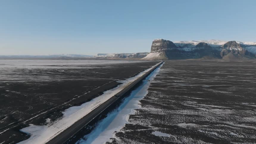 An aerial view follows a single dark car driving along a straight road through a vast snowdusted rural landscape towards distant cliffs under a clear sky