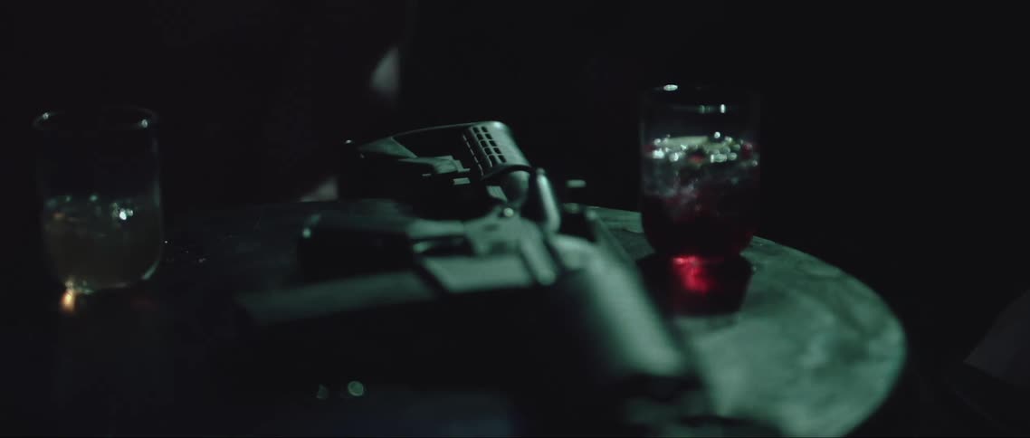 A dimly lit room features a table with an automatic rifle placed between two glasses of drinks