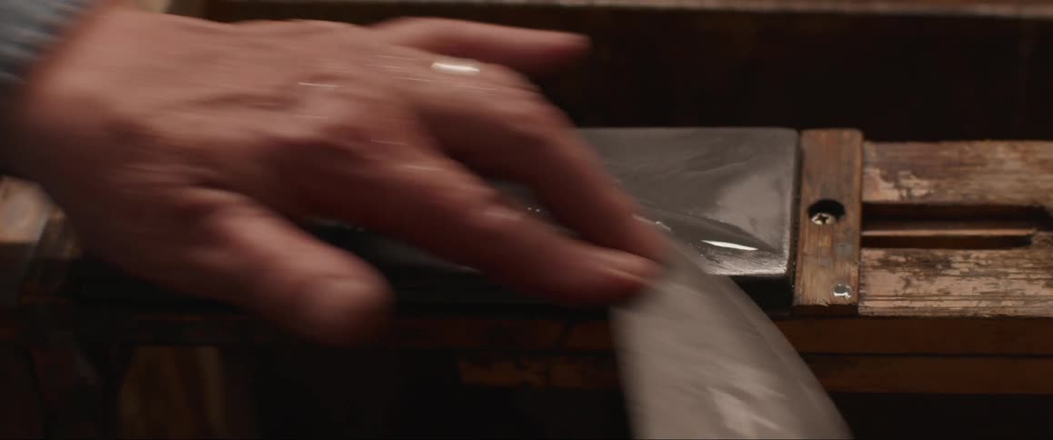 A hand sharpens a knife on a whetstone secured to a wooden base
