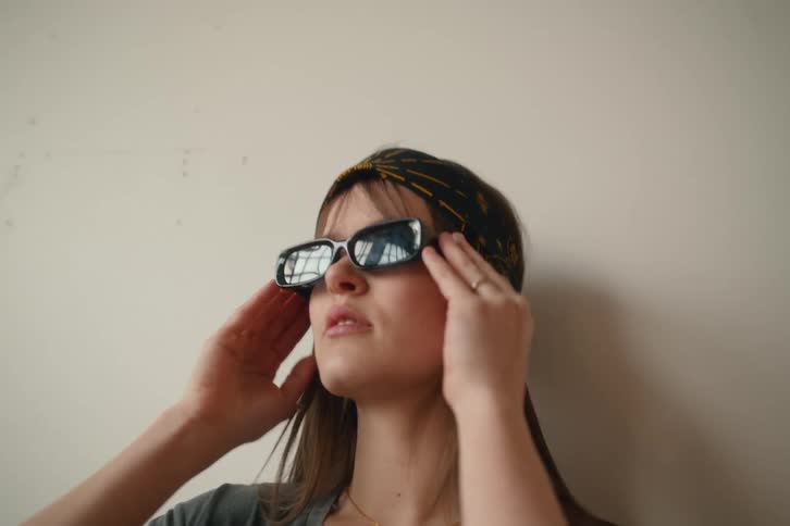 A young woman with long brown hair wearing a patterned head covering and sunglasses looks upwards against a plain light colored background