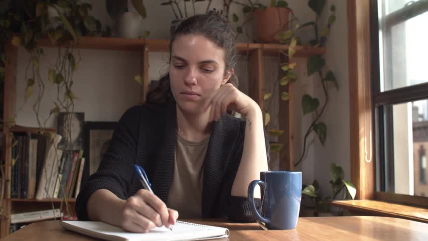 A woman with curly dark hair wearing a dark cardigan sits at a wooden table writing in a notebook with a blue pen next to a blue mug