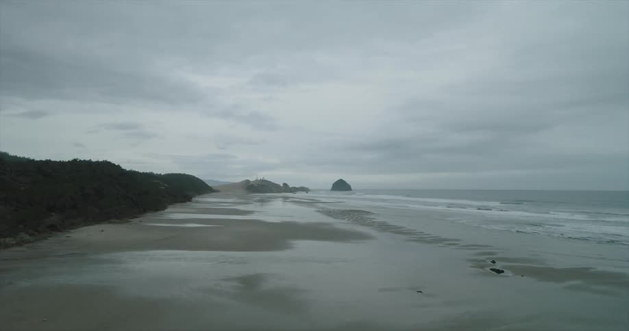 A wide overcast beach scene features a dark hillside wet sand reflecting the cloudy sky and the ocean stretching into the distance with a small rock formation visible