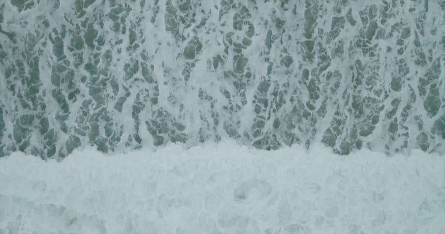 An aerial view of the swirling patterns of frothy waves crashing against each other in the ocean creating a dynamic texture across the surface