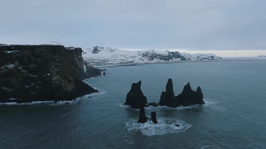 A shot from the air of an arctic coastal scene focused on stones in the ocean with snow capped mountains in the background and a black cliff in the foreground