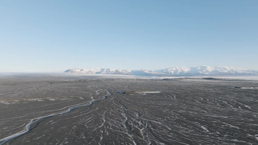 A vast desolate terrain features winding streams and snowcapped mountains under a clear blue sky