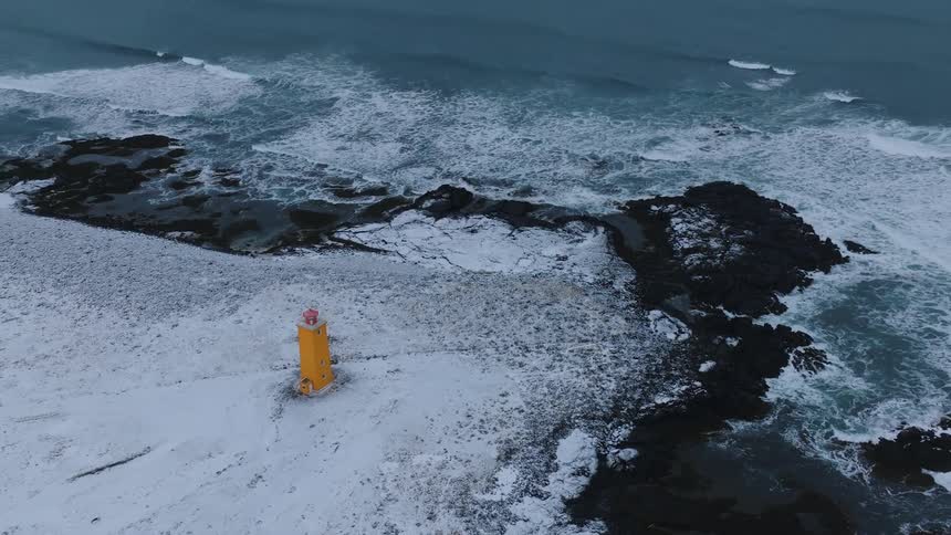A yellow lighthouse with a red top stands on a snowcovered rocky coastline as waves crash against the shore under a dim sky