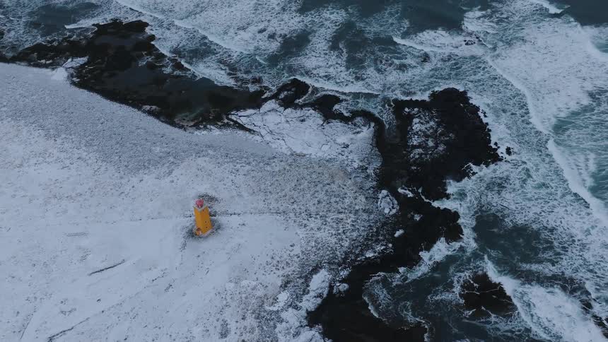 From a high angle a solitary yellow lighthouse stands on a snow covered landscape bordering a turbulent dark ocean with whitecapped waves crashing against rocky shores