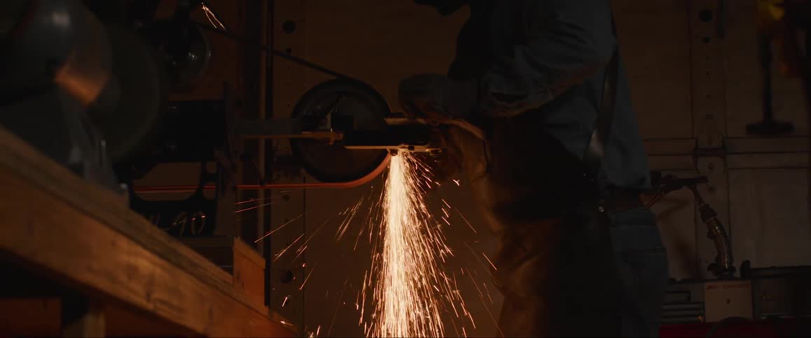A blue collar factory worker operates a metal grinder inside of an industrial warehouse
