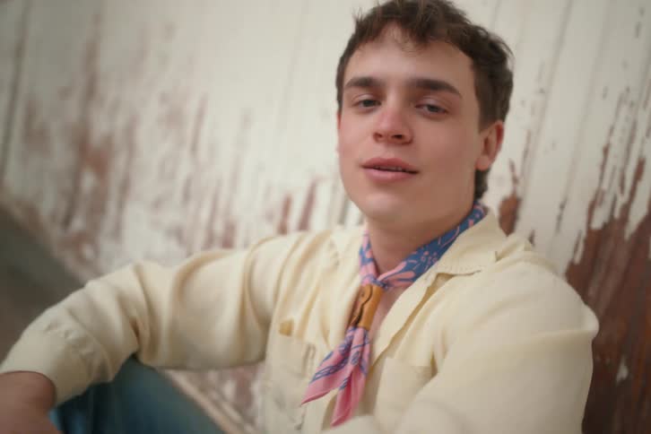 A young man with dark hair wearing a light colored shirt and a patterned neckerchief sits on a weathered surface and looks off to the side with a contemplative expression