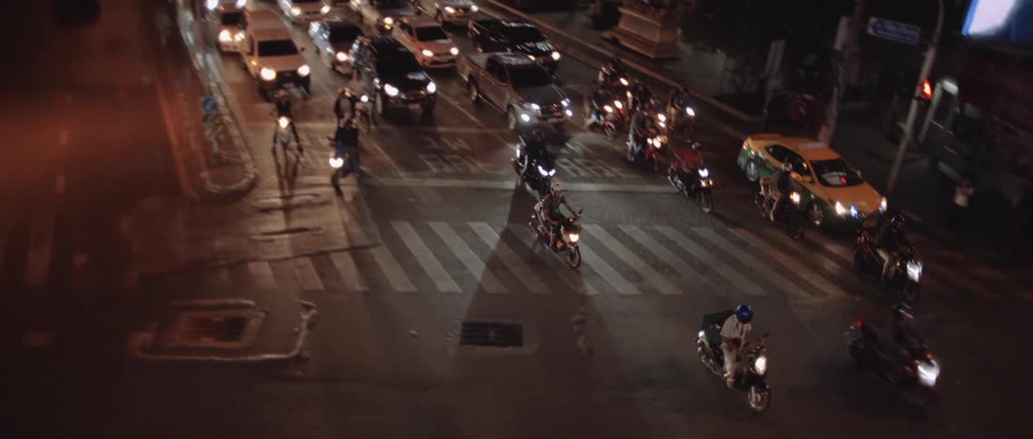 At night a busy city street shows cars and numerous motorcycles moving through an intersection with illuminated buildings in the background