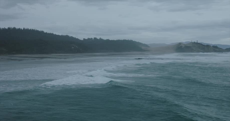 A wide overcast coastal scene captures a grey turbulent ocean with whitecapped waves leading to a dark forested hillside and a rocky outcrop in the distance under a cloudy sky