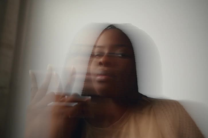 A person with long dark hair and brown skin laughs with their eyes closed raising a hand in a victory or peace sign