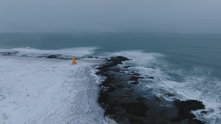 A snow covered shoreline meeting a turbulent sea with a solitary yellow structure standing out against the monochrome landscape