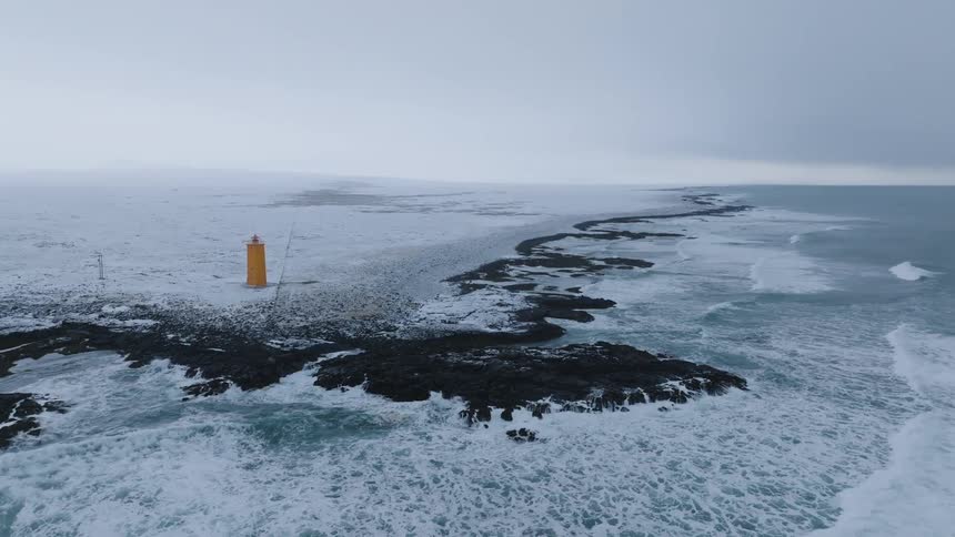 An aerial shot depicts a remote snow covered landscape where a lighthouse stands on a dark rocky outcrop amidst crashing white waves and a vast expanse of gray overcast sky