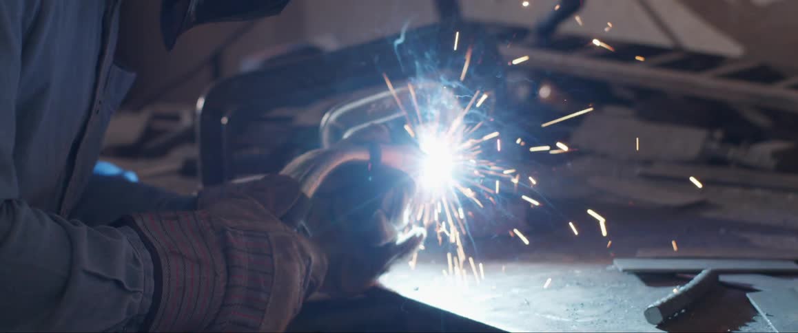 A person wearing protective gear is welding metal producing bright sparks and smoke