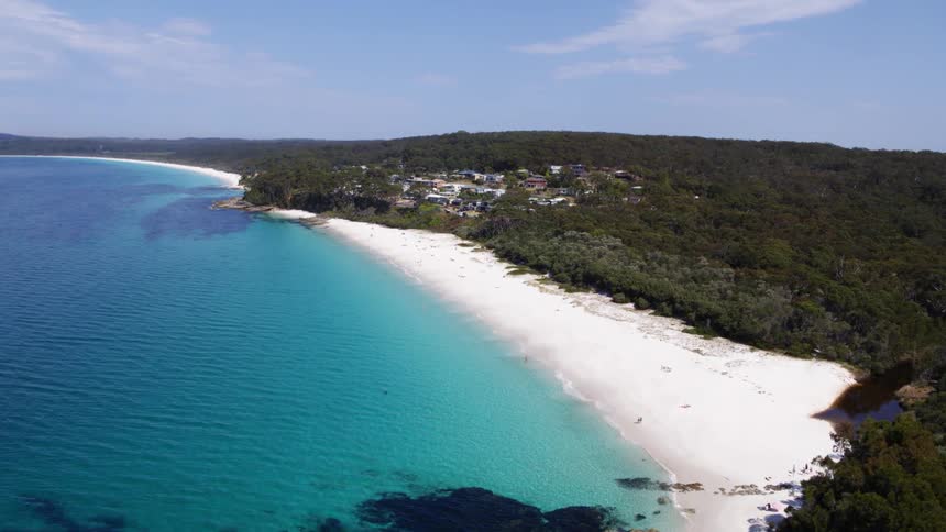 An aerial view of a stunning coastline with a white sandy beach meeting turquoise waters bordered by lush green forest and a small settlement