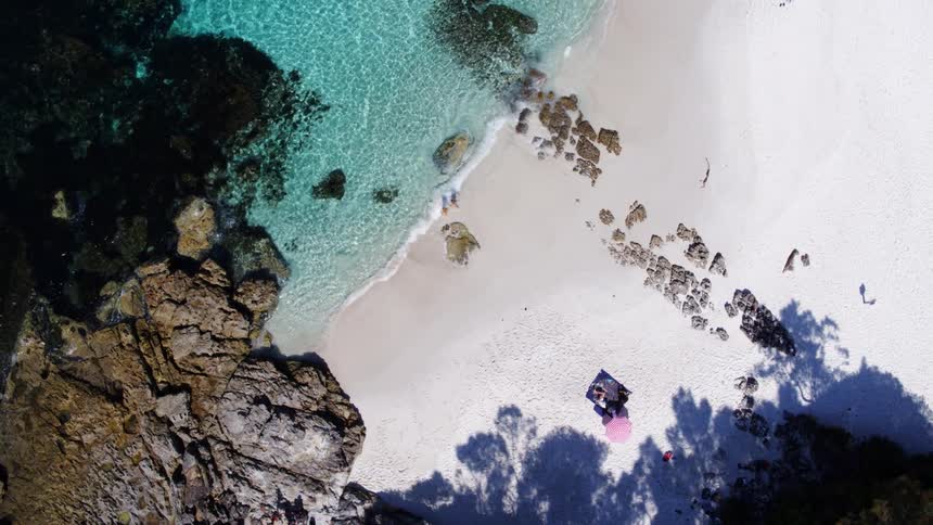 From a high angle a white sandy beach meets clear turquoise water with scattered rocks where a few people are relaxing under a striped umbrella and walking along the shore