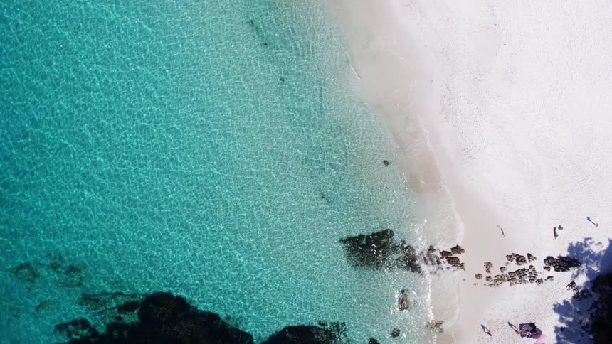 A pristine white sandy beach meeting clear turquoise water with two small figures visible on the sand creating a serene and picturesque coastal scene