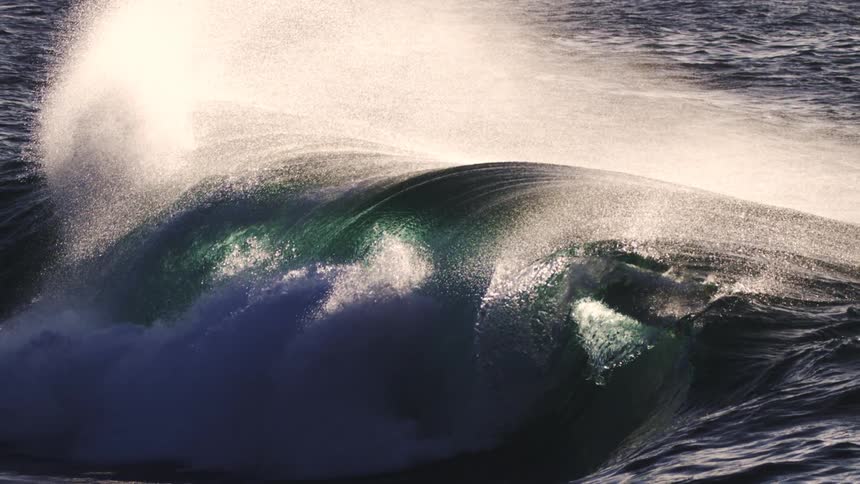 A large wave its water a deep green curls and crashes sending up a burst of white spray against the darker ocean surface
