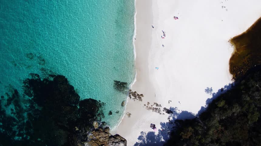 An aerial view of the stunning contrast of a white sandy beach meeting turquoise water with dark rocks visible at the waters edge and small figures dotting the sand