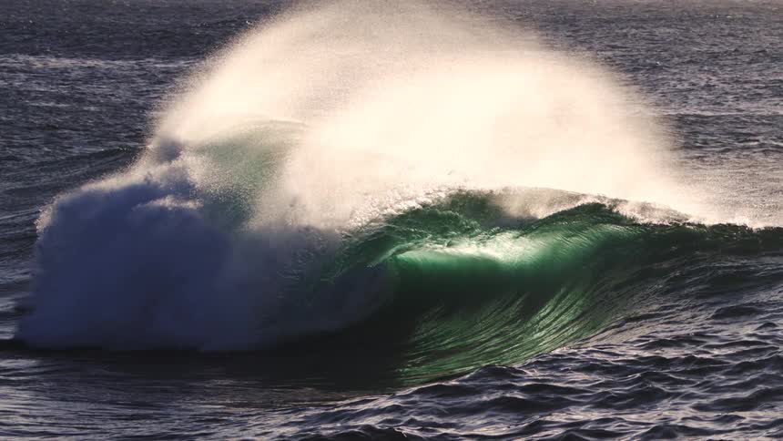 A dark rolling wave its peak catching a glint of sunlight moves across the textured surface of the ocean