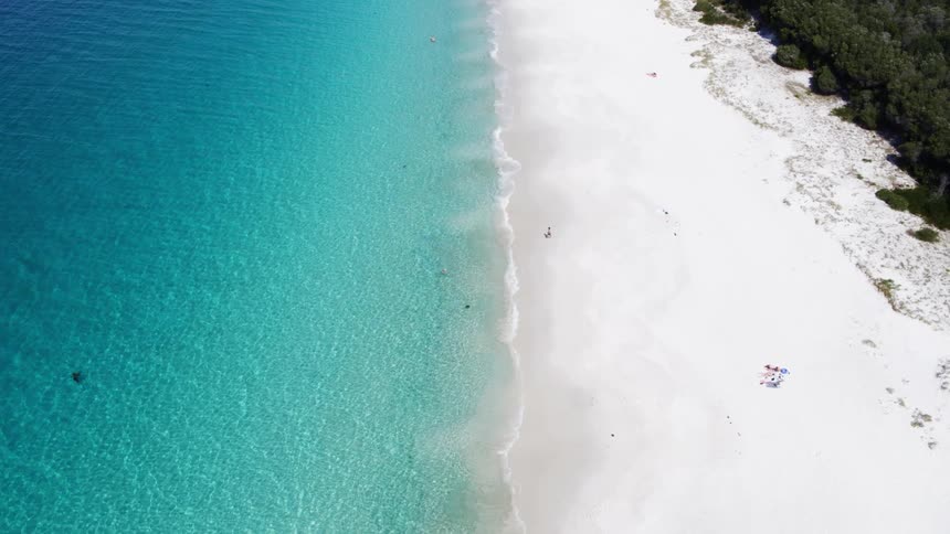 A person is walking across white sand toward crystal blue water of the sea