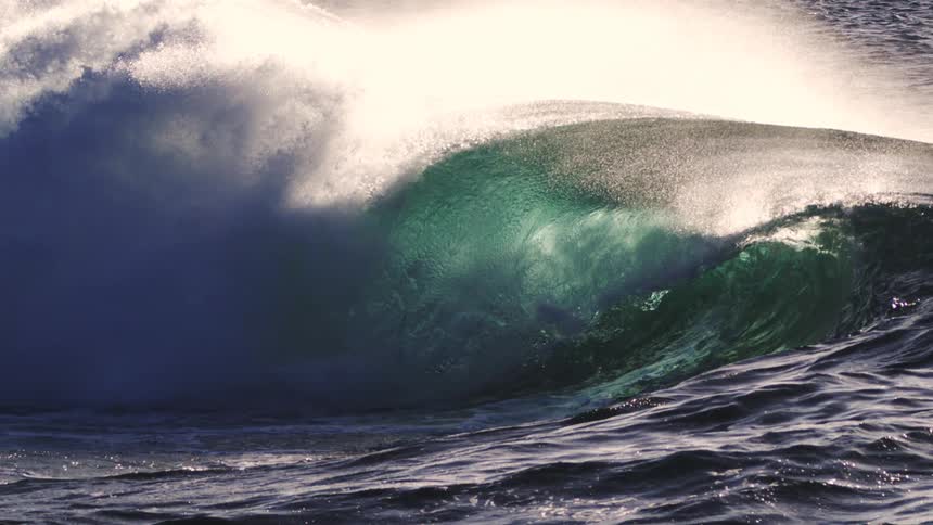 A massive powerful wave crests dramatically against the sunlight sending a spray of water into the air