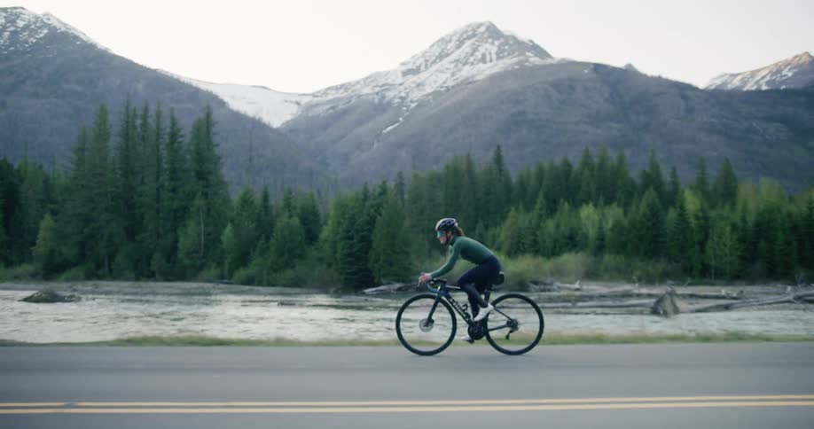 A cyclist rides along an asphalt road with a flowing river on one side and a dense forest leading to snow capped mountains in the background