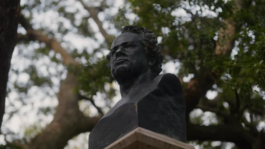 A darkcolored bust of a person with curly hair is mounted on a pedestal framed by a backdrop of tree branches and leaves