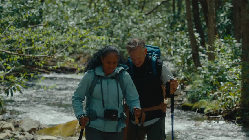 Two backpackers with hiking poles stand facing each other near a flowing stream in a wooded area