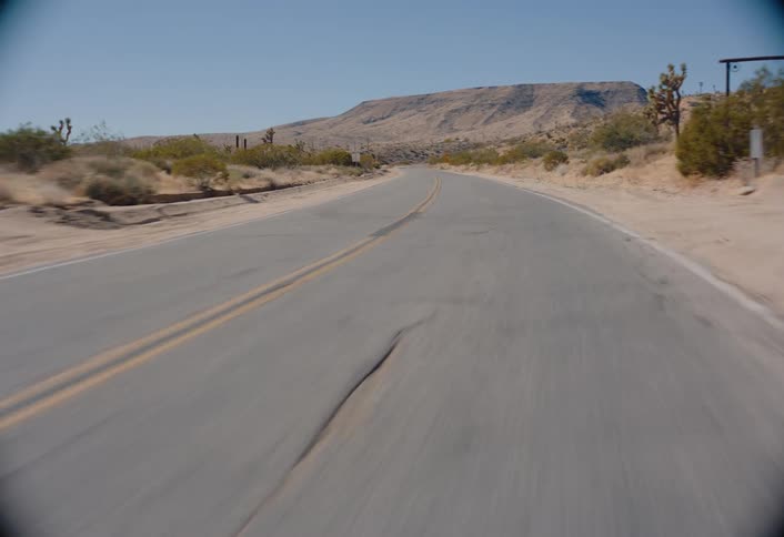 An empty asphalt road stretching into the distance through a desert landscape with dry shrubs and a rocky hill visible under a clear blue sky