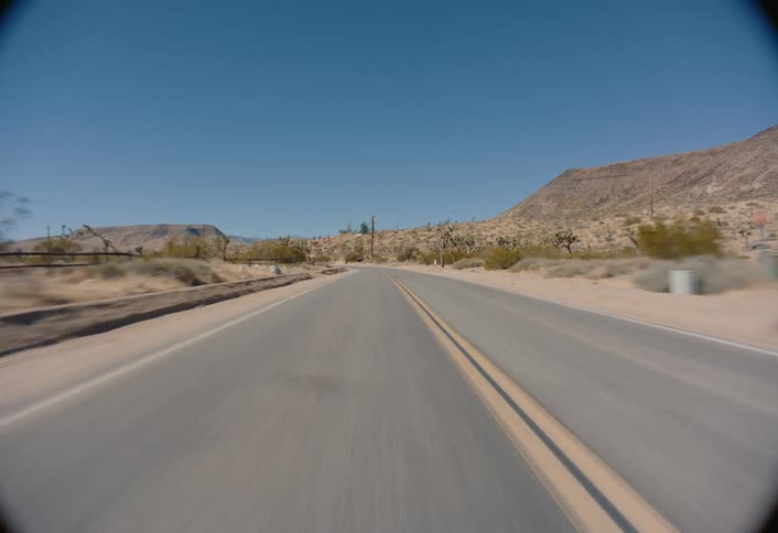 A moving vehicle captures an asphalt road stretching into the distance through a desert landscape with dry shrubs and a rocky hill visible under a clear blue sky