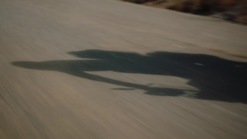 An asphalt highway flies by as a motorcycle catches up to the camera casting a full shadow on the highway of the bike and rider as they speed by