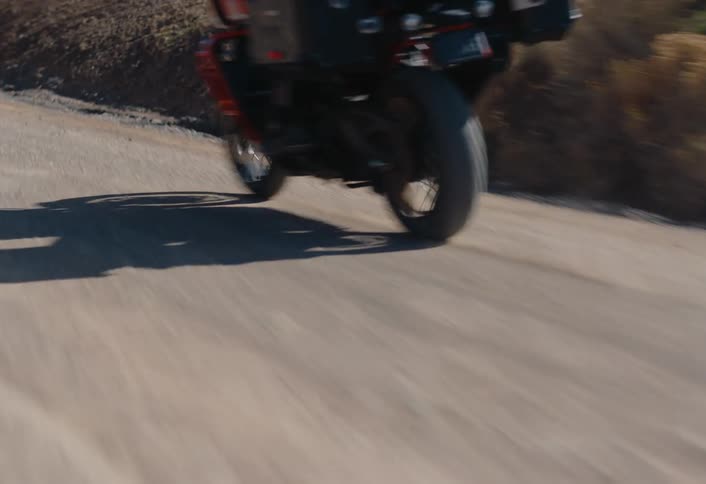 A moped rider swereving back and forth on a gravel road through dry foothills