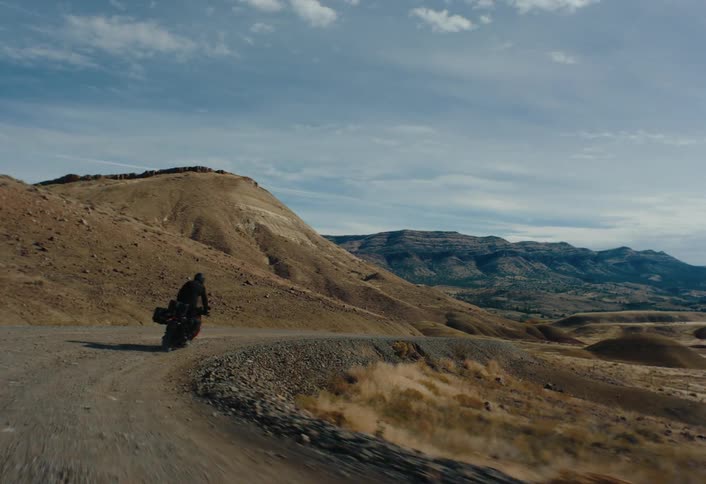 A lone figure on a motorcycle travels along a winding dirt road through a hilly arid landscape under a partly cloudy sky
