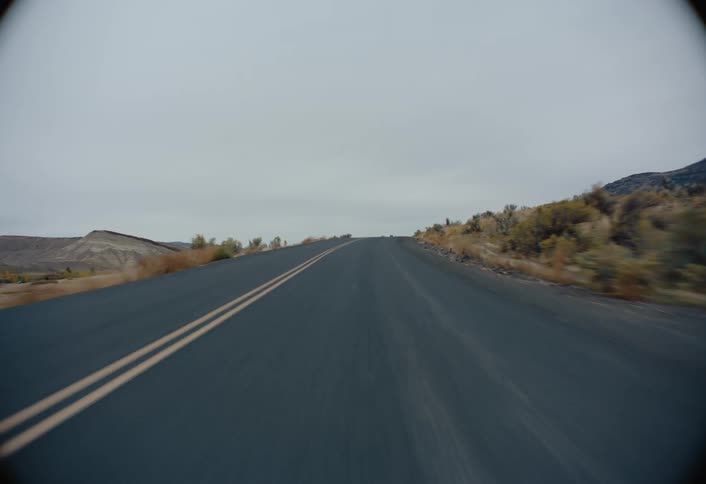 A moving vehicle captures an asphalt road stretching into the distance through a desert landscape under an overcast sky with dry shrubs lining the sides and hills visible in the background