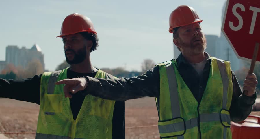 Two construction workers wearing hard hats and safety vests are positioned near a construction site holding a stop sign and gesturing with his arm extended