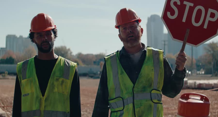 Two construction workers in hard hats and safety vests stand in a construction zone one holding a stop sign