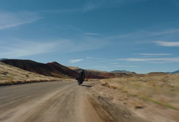 A lone motorcyclist travels down a straight sunlit road that cuts through a vast arid landscape of rolling hills under a bright blue sky